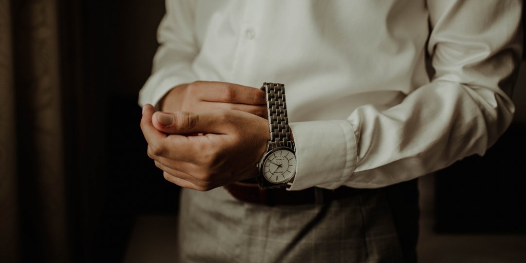 Mężczyzna ubrany w białą koszulę i szare spodnie, zakładający srebrny zegarek na nadgarstek.
