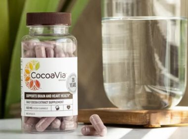 Butelka kapsułek CocoaVia obok szklanki wody, na stole przed zielonymi roślinami doniczkowymi.