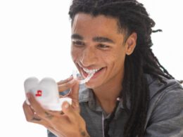 Młody mężczyzna uśmiechający się i trzymający skrzynkę ustalającą Bajt podczas umieszczania bajtowego Clear aligner na zębach.