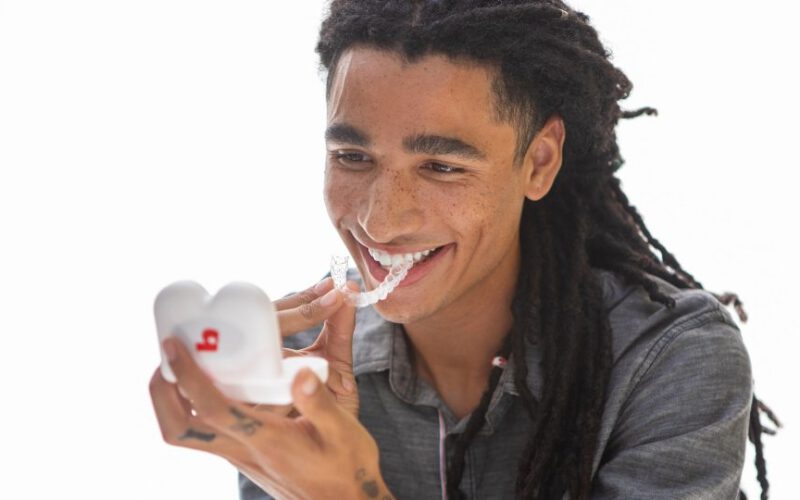 Młody mężczyzna uśmiechający się i trzymający skrzynkę ustalającą Bajt podczas umieszczania bajtowego Clear aligner na zębach.