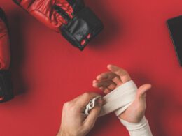 ujęcie boksera zakrywającego ręce w elastyczny bandaż na czerwonej powierzchni rękawicami bokserskimi i smartfonem oraz ciężarami