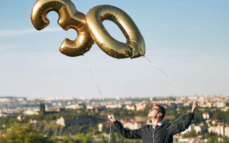 Mężczyzna trzymający balony helowe w kształcie numer 30 przeciwko miastu.